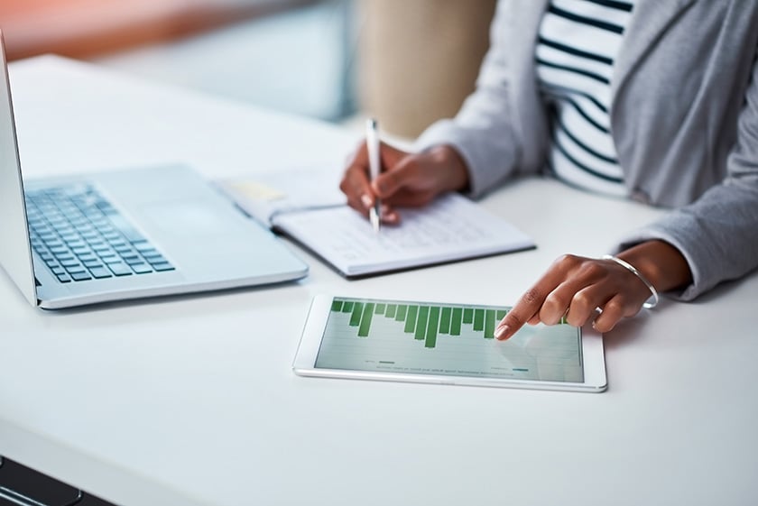 Женщина переписывает финансовую информацию с диаграммы на планшете и стоящего перед ней ноутбука