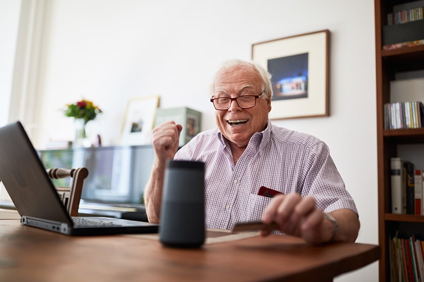  Пожилой мужчина делает покупки в Интернете с помощью кредитной карты, ноутбука и &quot;умной&quot; колонки.